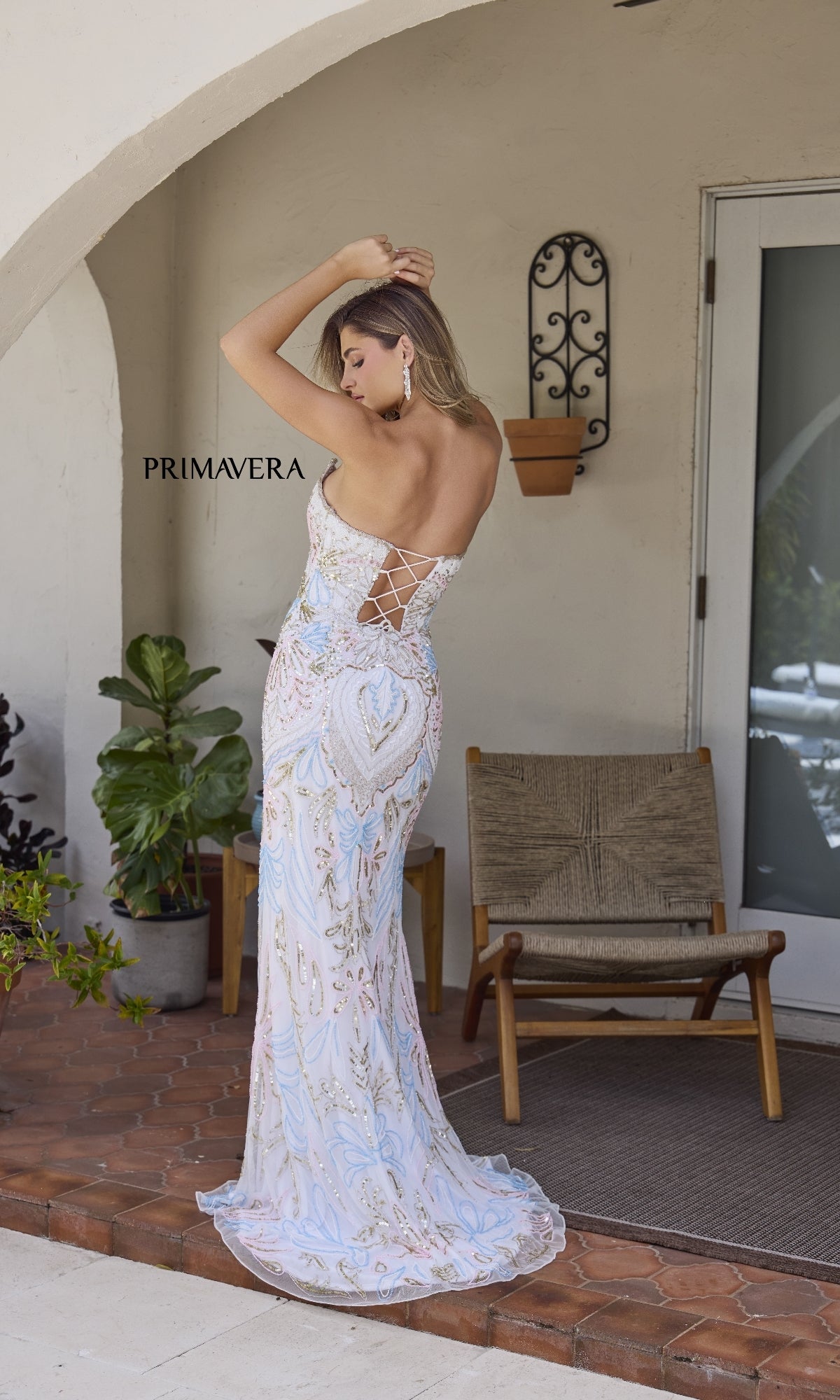 A woman wearing the Strapless Long Beaded Prom Dress: Primavera 4382 with a lace-up back stands on a tiled patio near a chair, lush plants, and a glass door.
